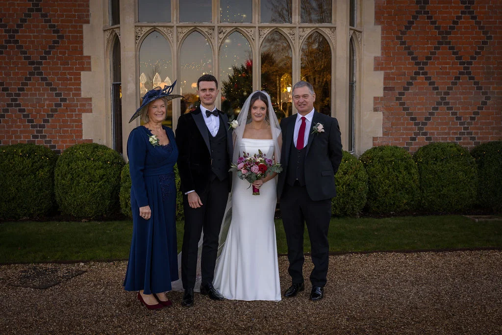 A bridal party including the bride and groom alongside the mother and father of the bride poses outdoors in front of a grand brick manor house with large arched windows.