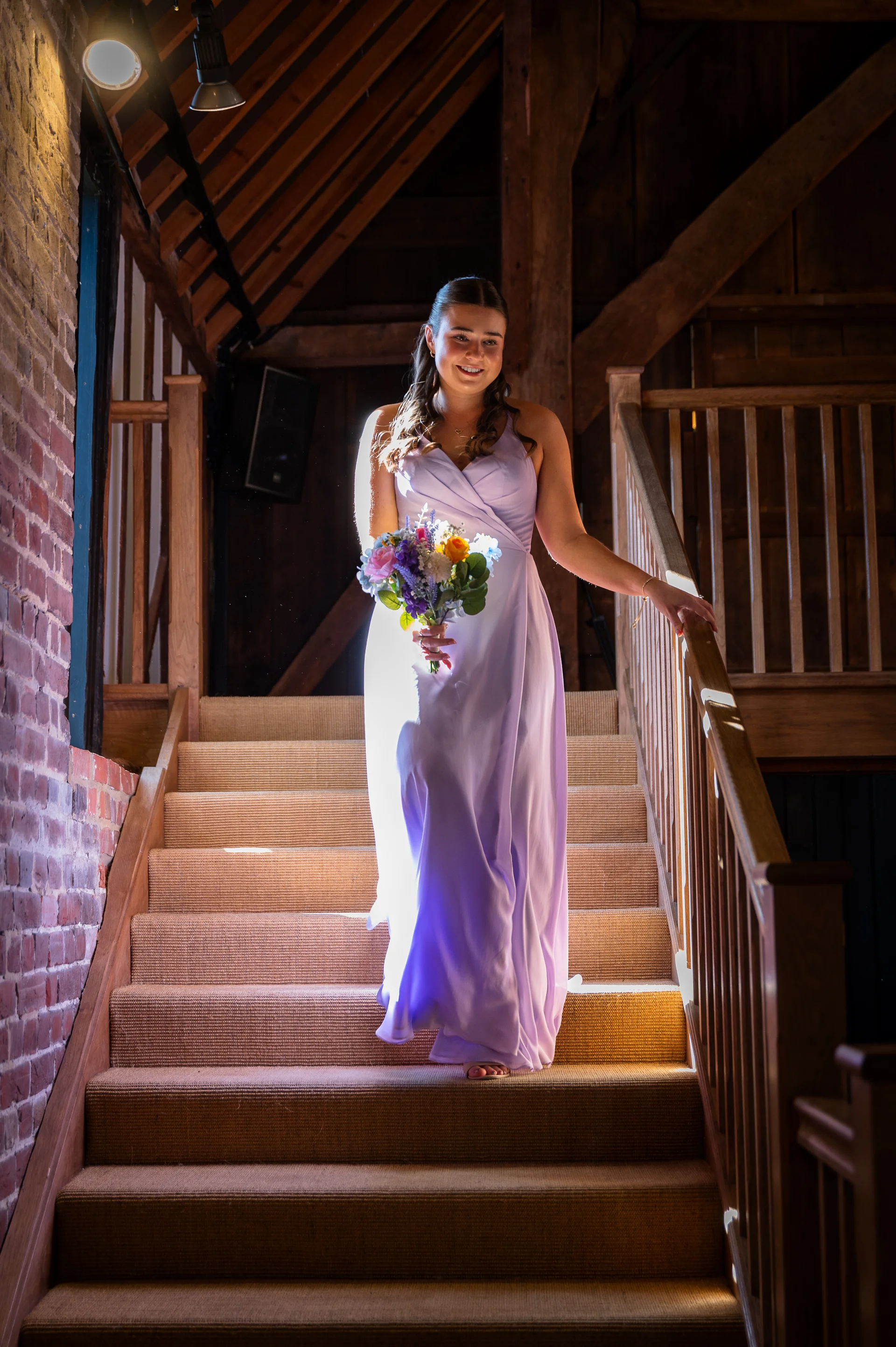 A bridesmaid in a light purple dress smiles while walking down a wooden staircase inside a rustic barn, holding a colourful bouquet of flowers.