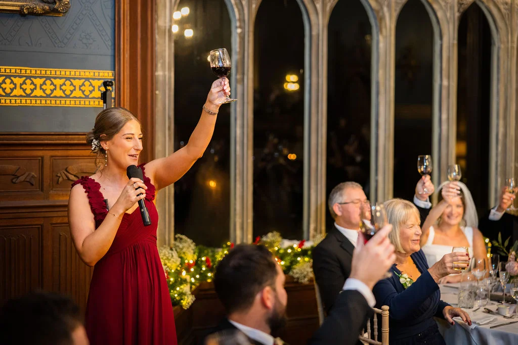 A bridesmaid in a burgundy dress delivers a speech at a wedding reception, holding a glass of wine aloft while guests raise their glasses in a toast.