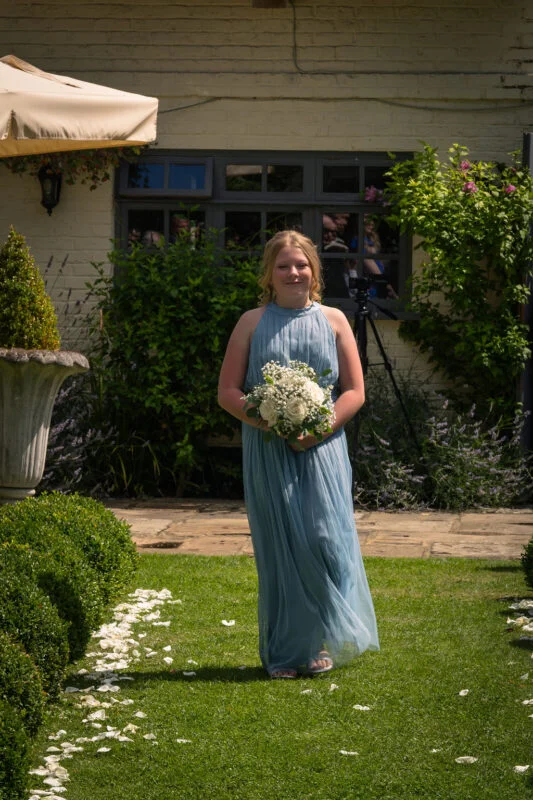 A young bridesmaid in a flowing dusty blue halter neck dress walks down a green lawn strewn with white petals, holding a posy bouquet of roses and baby's breath.