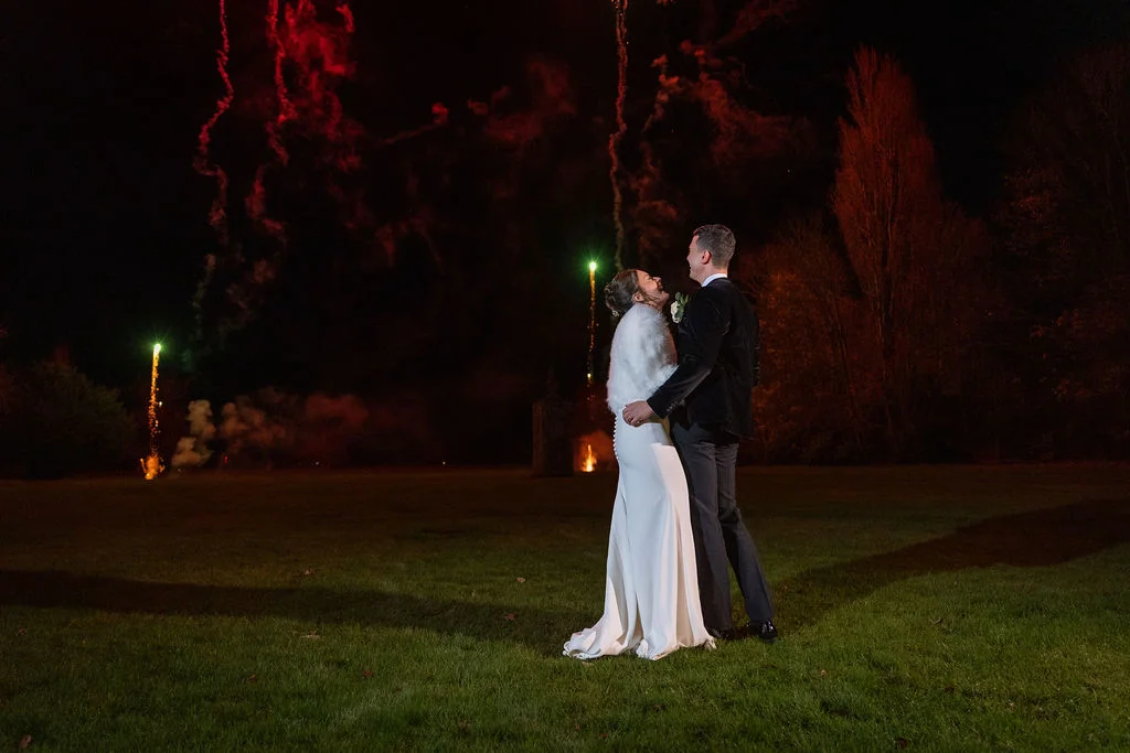 Newlyweds embrace on a grassy lawn at night while vibrant fireworks light up the dark sky behind them.