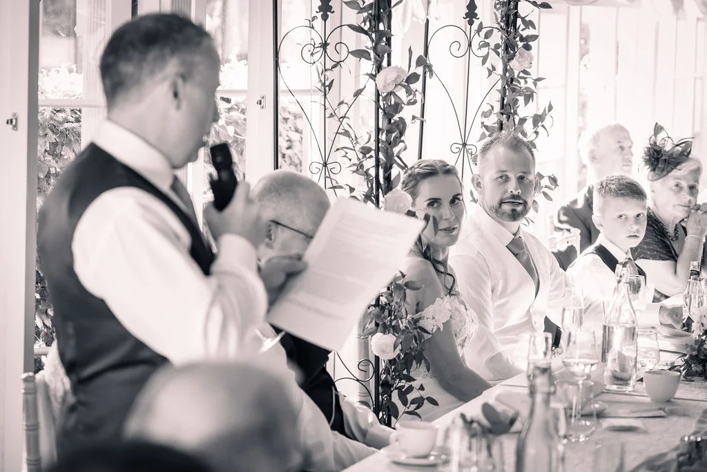 A wedding guest stands to deliver a speech while the bride and groom listen attentively from the top table during the reception.