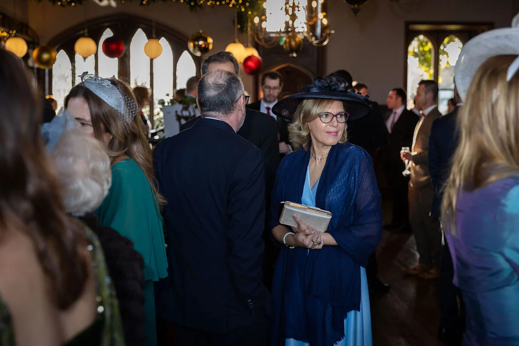 A festive winter wedding reception where guests mingle inside a grand venue adorned with seasonal decorations and large windows.