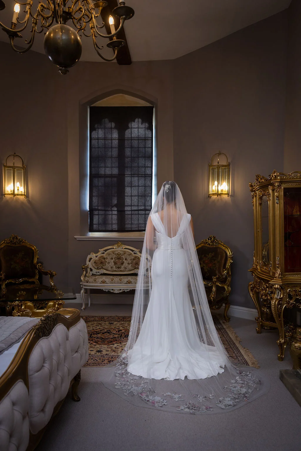 A bride stands in a grand, classically styled room facing away from the camera, wearing an elegant white crepe gown and a long, intricately embroidered veil.