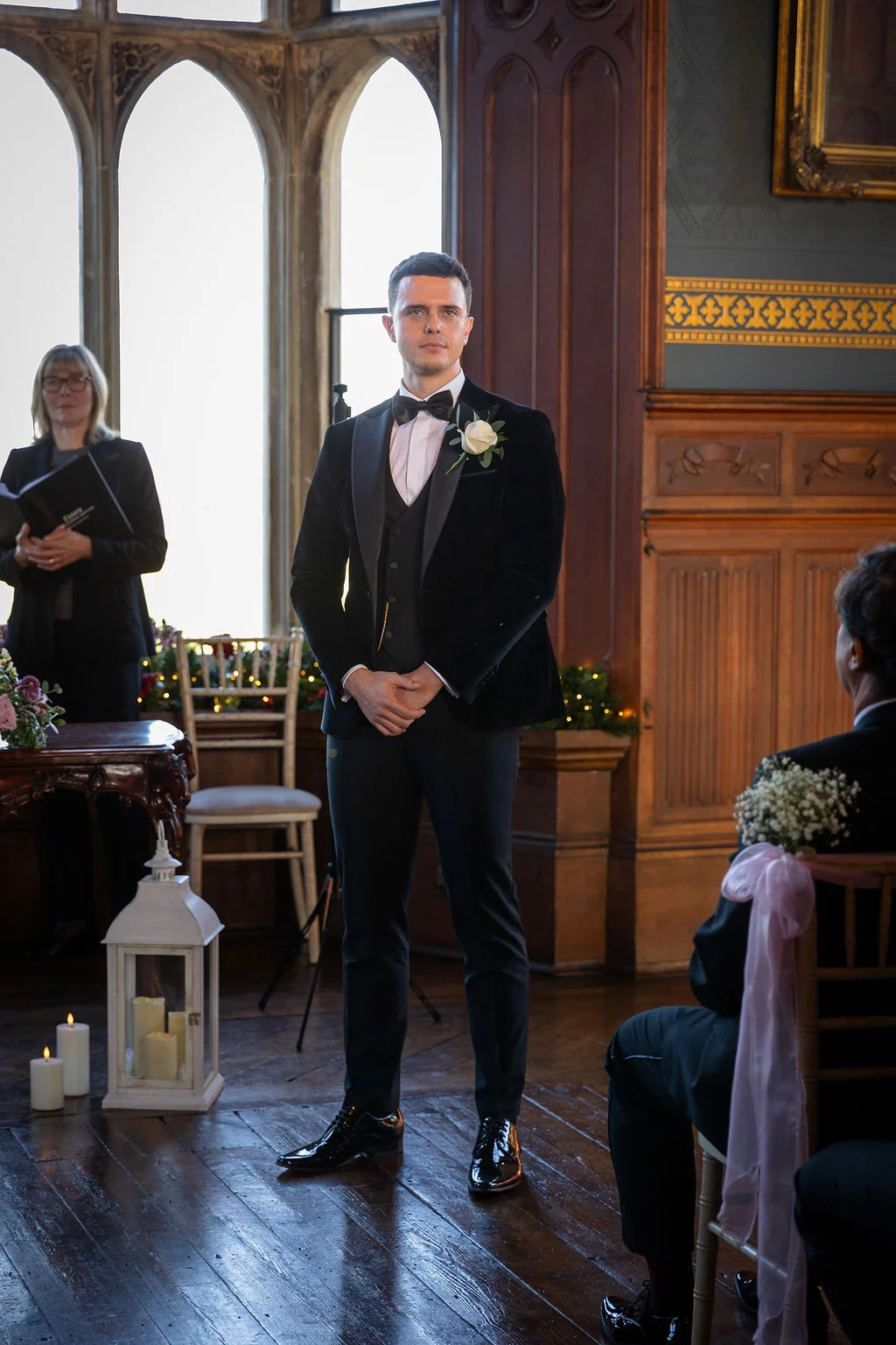 A handsome groom in a black velvet tuxedo waits calmly at the altar inside a historic panelled room with large arched windows during a wedding ceremony.