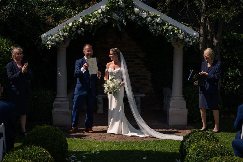 A newly married couple stands beneath a floral-decorated gazebo during an outdoor wedding ceremony, with the groom proudly holding up their signed marriage certificate.