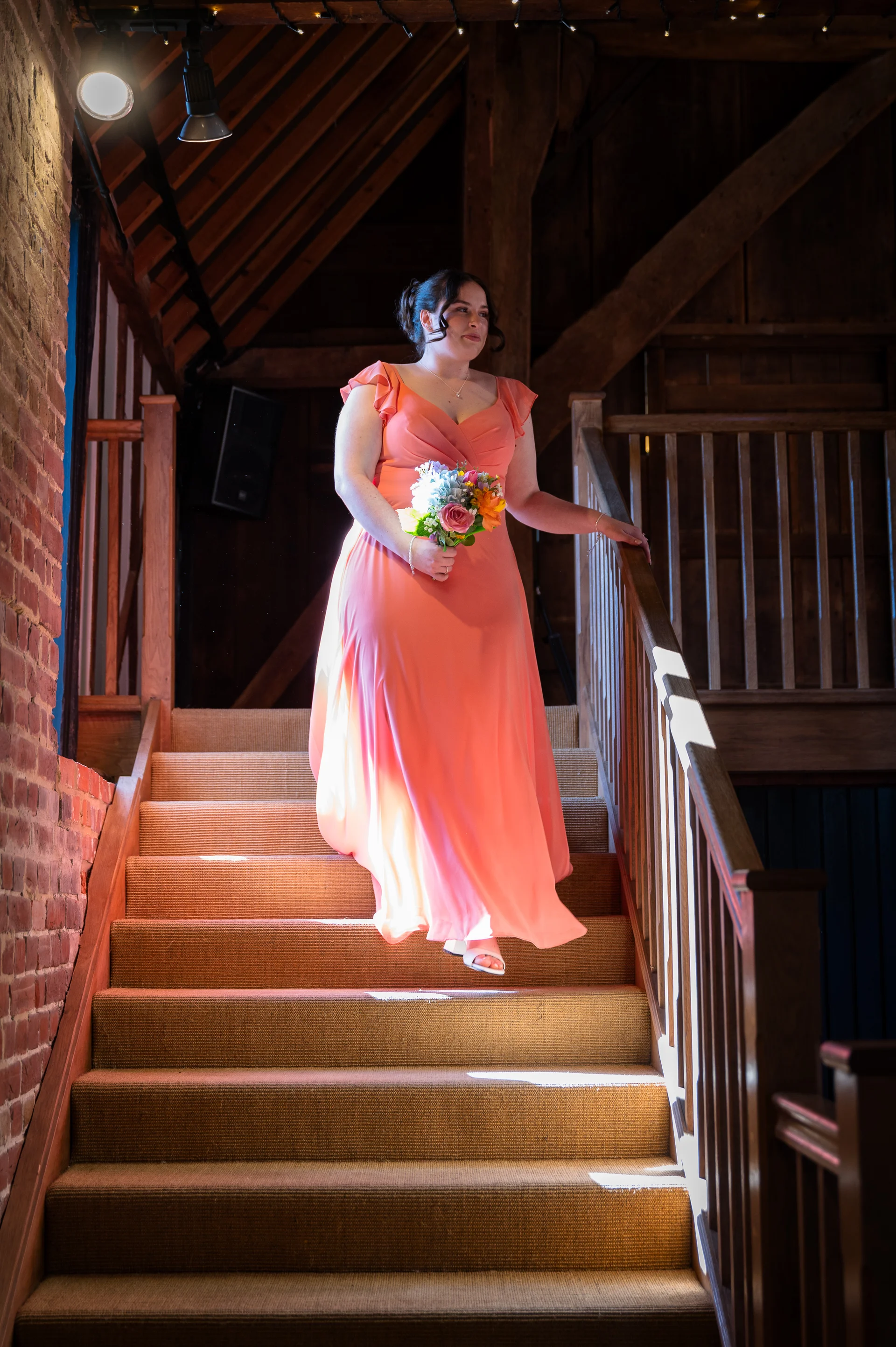 A smiling bride wearing a vibrant coral v-neck chiffon dress descends the carpeted wooden staircase of a rustic barn venue, holding a colourful bouquet of flowers.