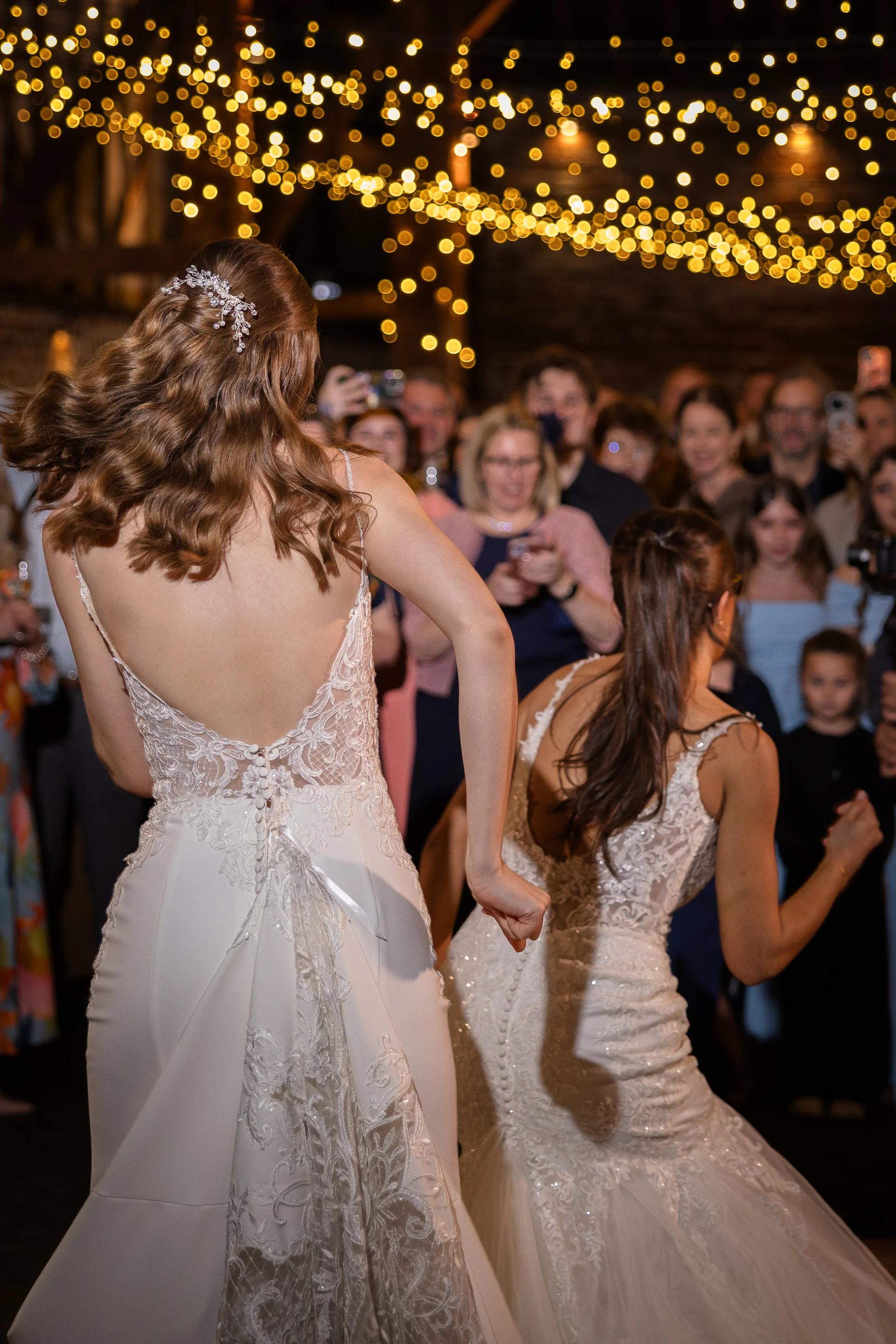 Two brides in stunning lace-detailed backless wedding dresses energetically dance together on the packed dance floor beneath a glowing canopy of fairy lights.