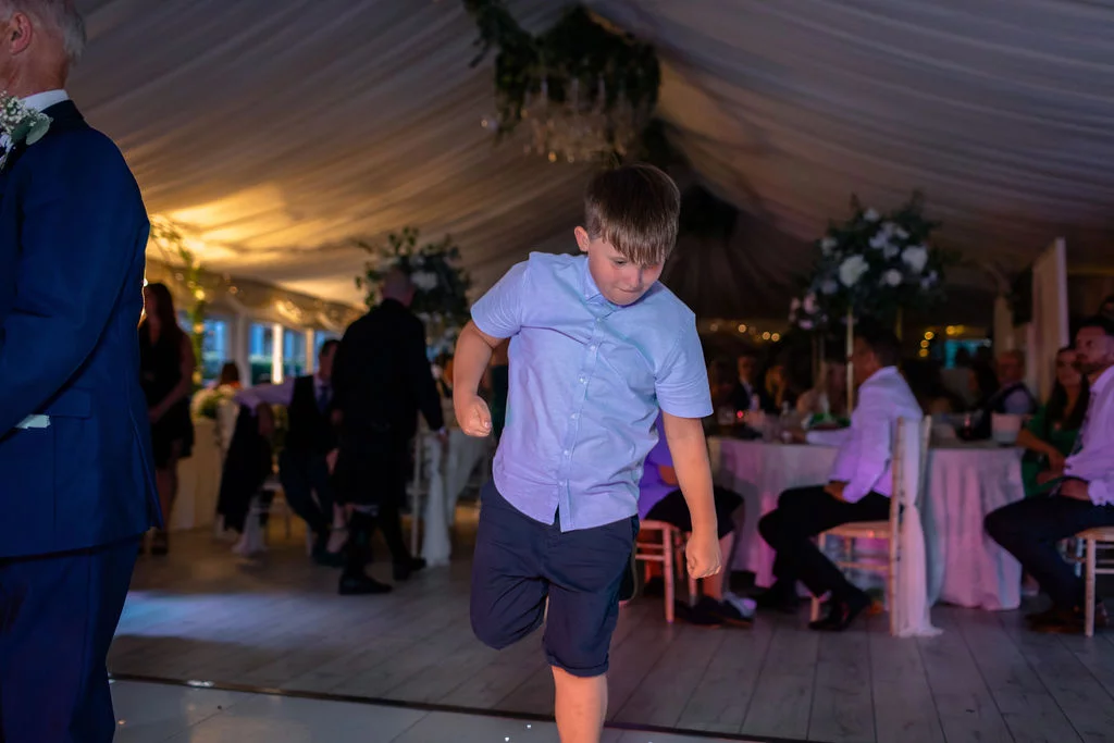 A young boy energetically dancing on the illuminated dance floor inside a marquee reception during the evening celebration.