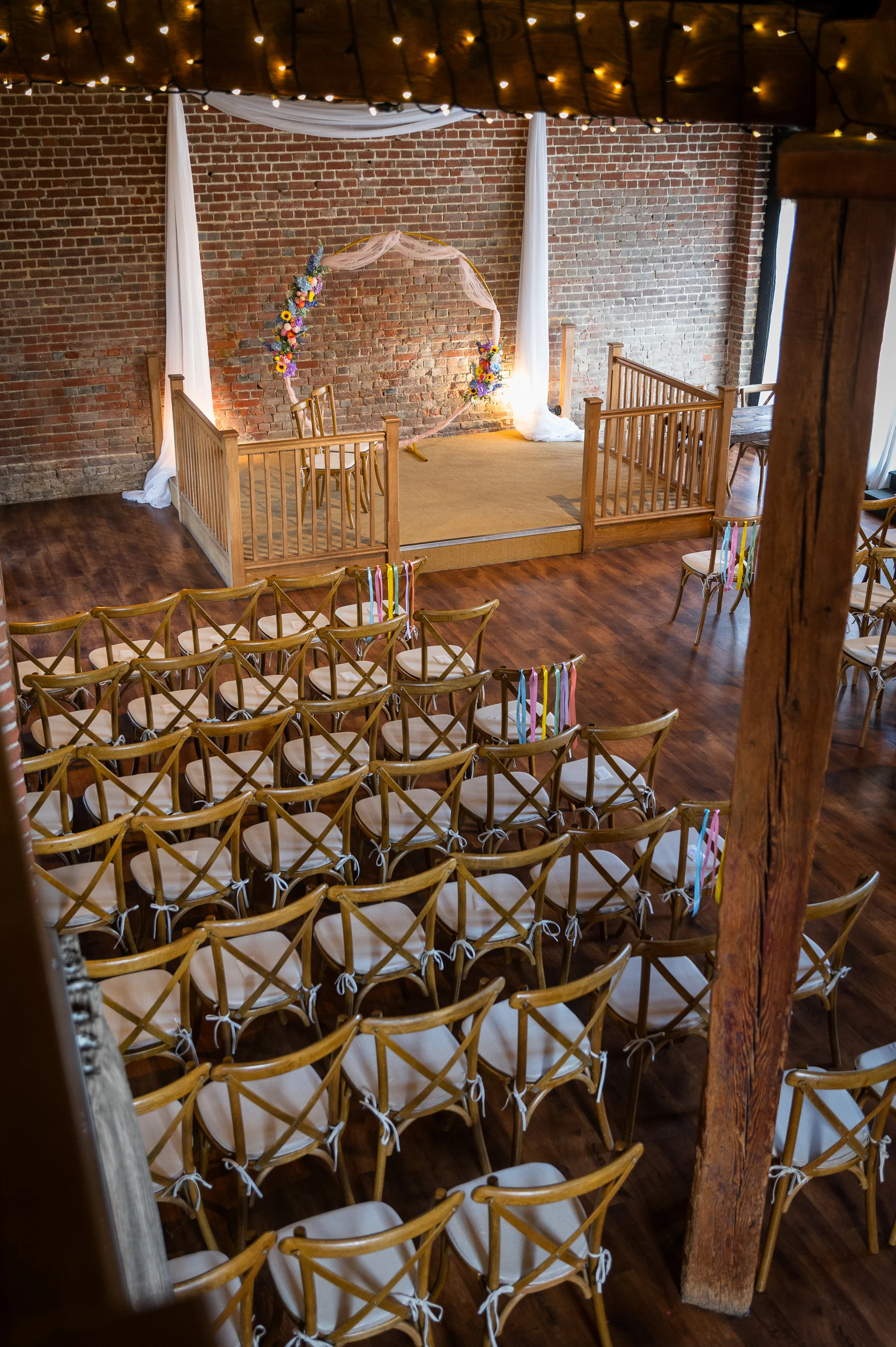 An overhead view of a rustic barn wedding ceremony venue featuring exposed brick walls, a decorated circular arch on a raised stage, and wooden cross back chairs adorned with colourful ribbons.