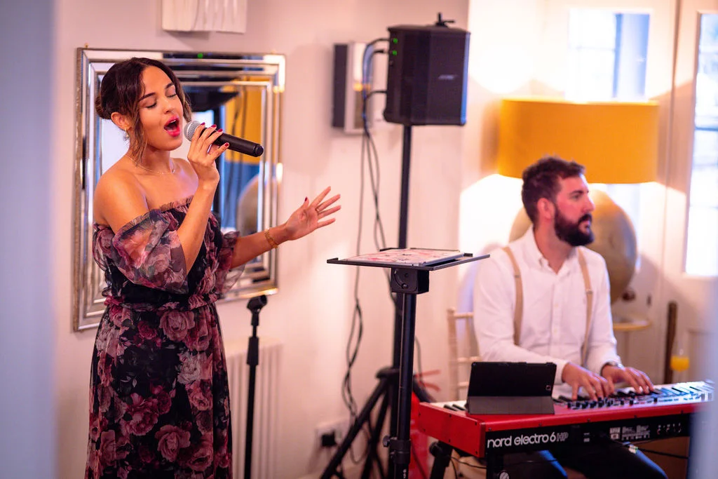 A female vocalist passionately sings into a microphone while performing alongside a keyboard player wearing braces during the wedding reception.