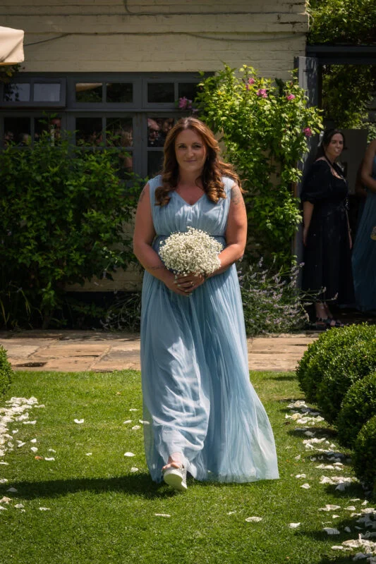 A bridesmaid in a flowing dusty blue gown walks down an outdoor aisle strewn with white petals, holding a bouquet of baby's breath.