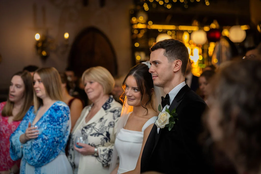 A newlywed couple smiling together amidst seated wedding guests in a warmly lit venue with a glowing bokeh background.