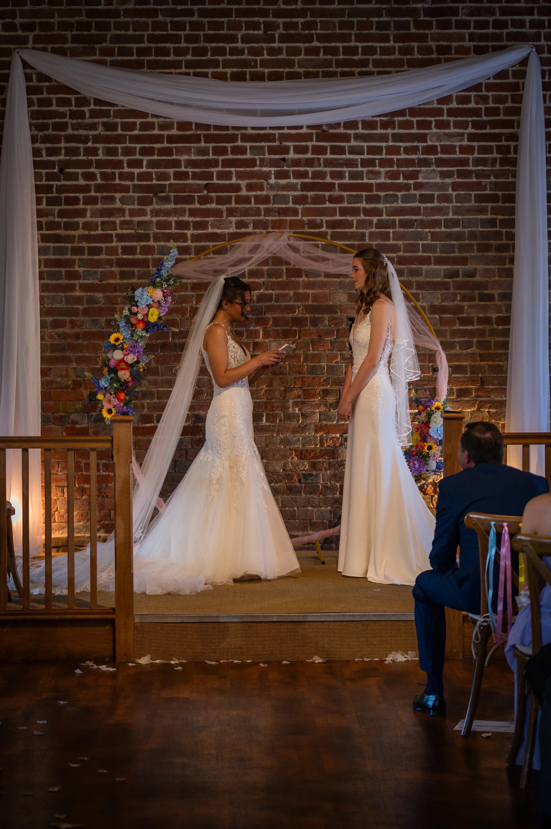 Two brides exchange personal vows during an indoor wedding ceremony before an exposed brick wall and a floral arch.