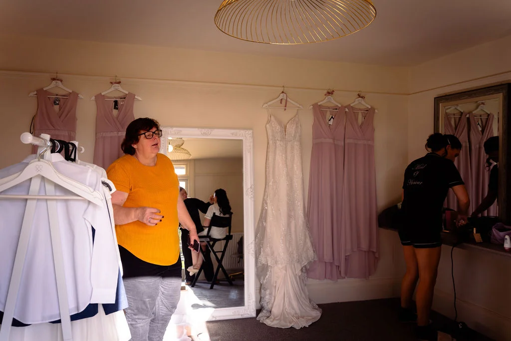 A bride and her bridal party prepare in a room decorated with hanging wedding dresses and pink bridesmaid gowns while a stylist applies makeup in the mirror.