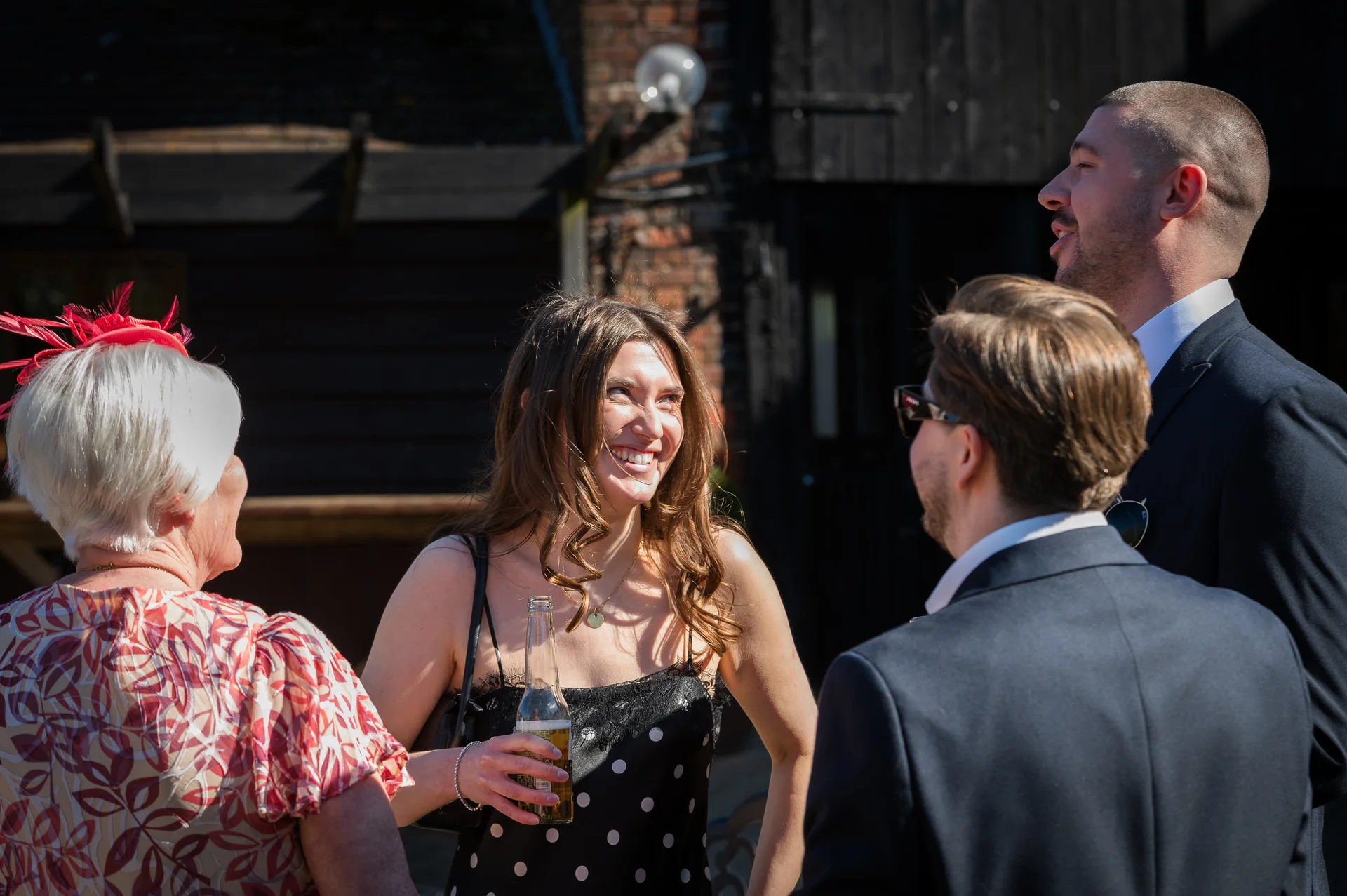 Wedding guests laugh and socialise outdoors in the sunshine during the drinks reception outside a rustic venue.