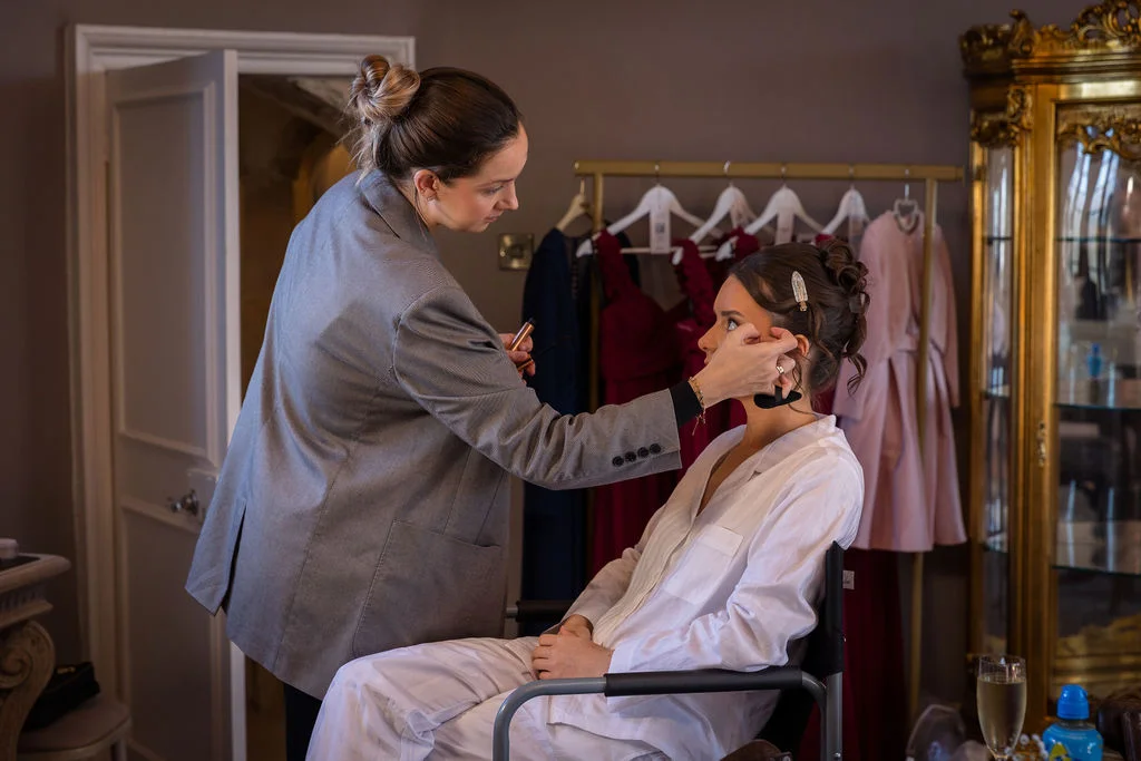 A professional makeup artist meticulously applies finishing touches to a bride sitting in a chair during morning preparations.
