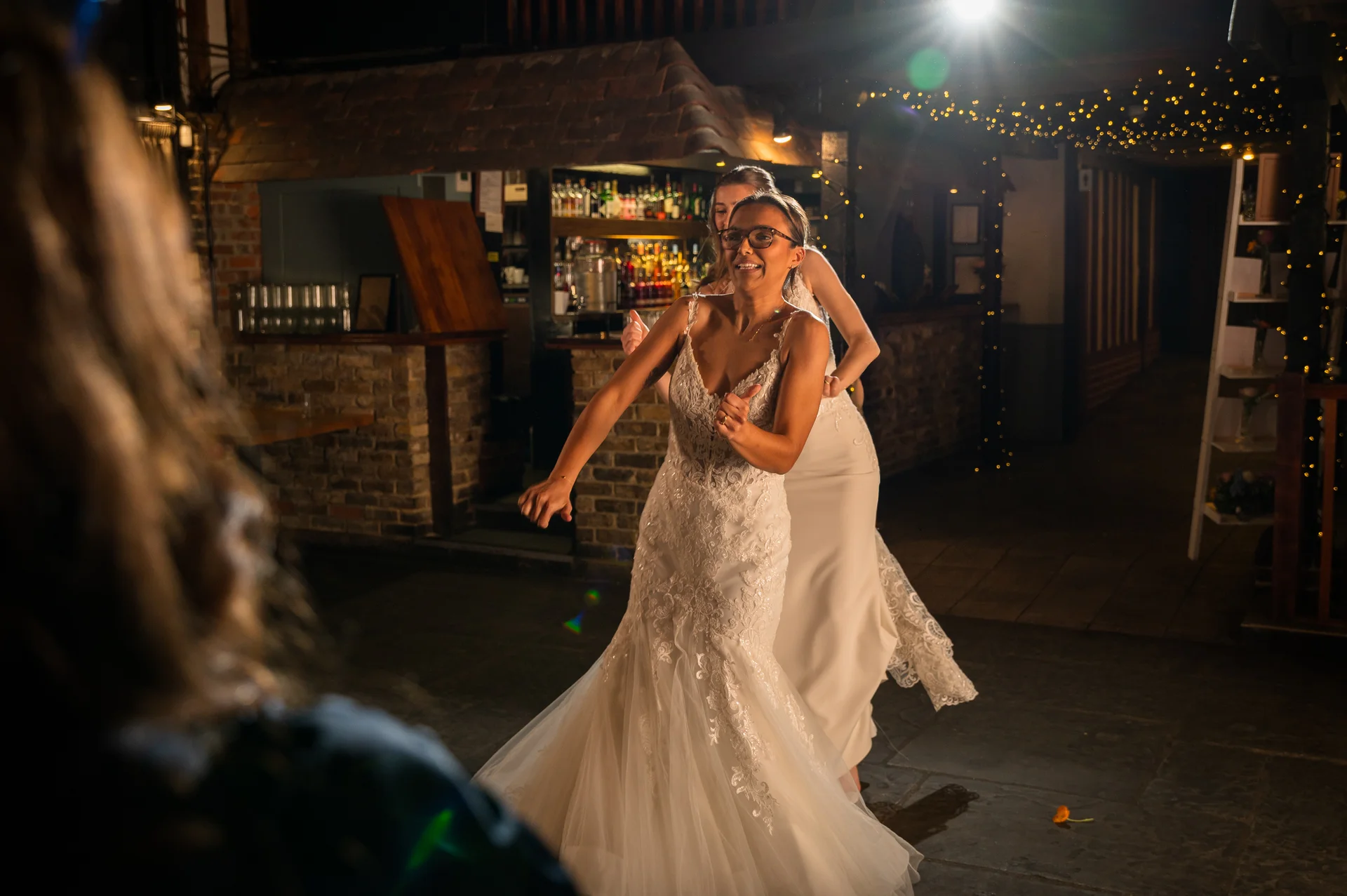 Two smiling brides in wedding gowns dance joyfully on an illuminated evening dance floor surrounded by fairy lights.
