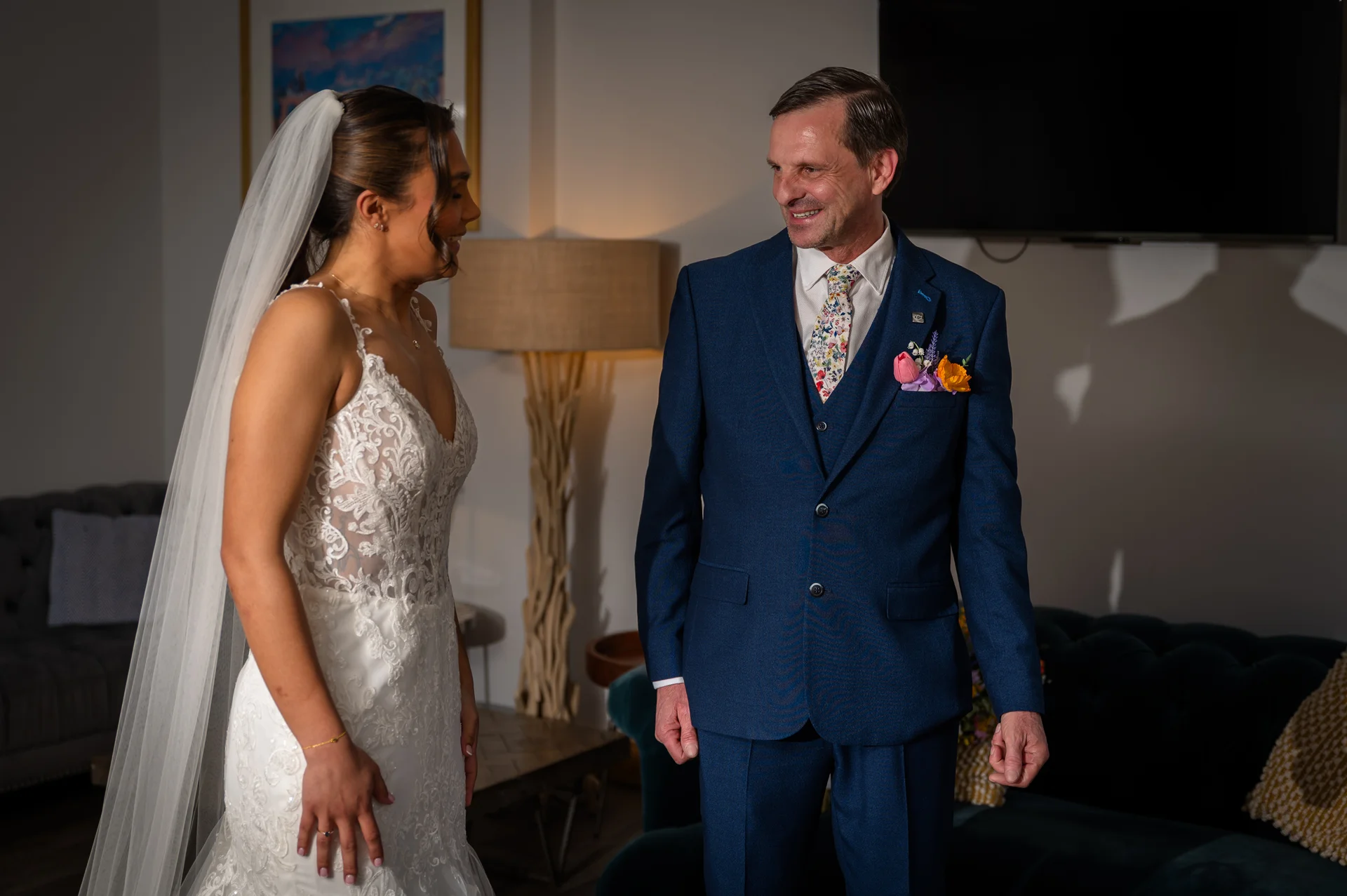 A smiling father in a blue suit lovingly looks at his daughter in a lace wedding dress and veil during a first look moment.