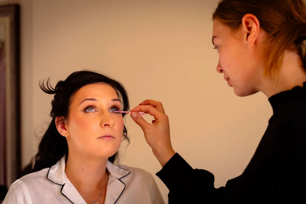 A makeup artist applies finishing touches to the bride's eyelashes with a pink wand while she sits calmly during bridal preparations.
