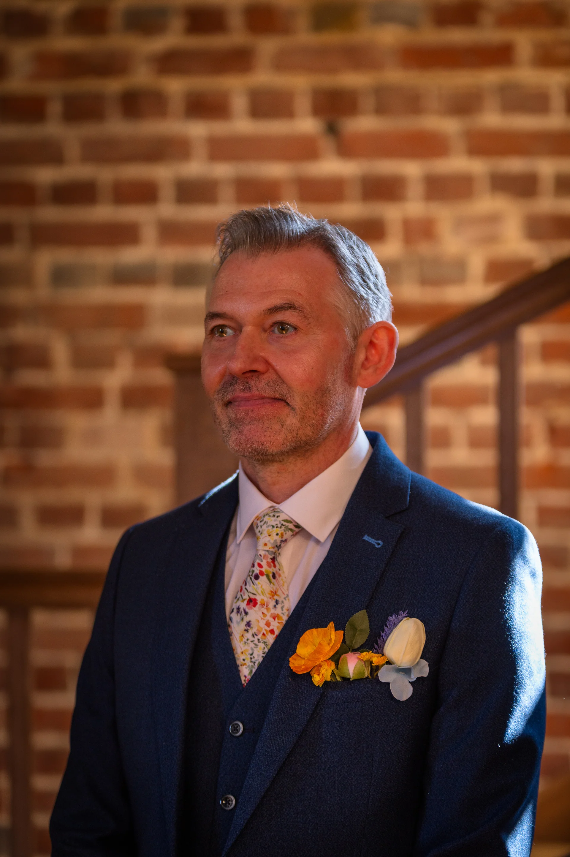 A smiling groom wearing a blue three-piece suit and a floral tie stands against an exposed brick wall interior.