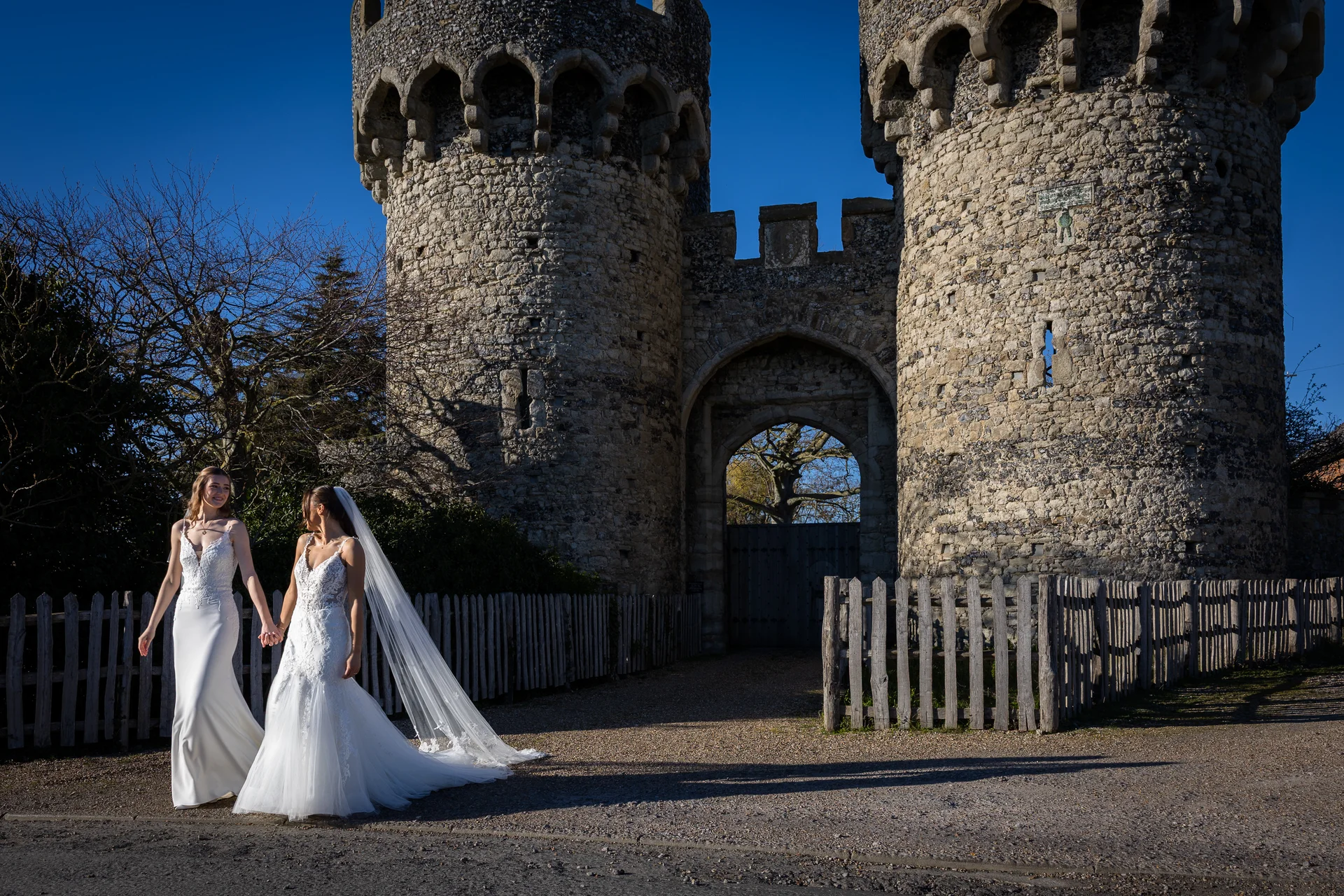 Two brides in wedding dresses holding hands while walking outside in front of an ancient stone castle under a bright blue sky.