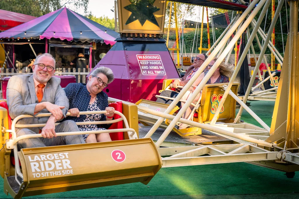 An older couple enjoys a playful moment riding a vintage fairground attraction at a festival-themed wedding reception.