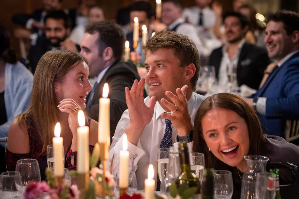 Wedding guests enjoy animated conversation and laughter during the wedding breakfast, surrounded by a candlelit table setting.