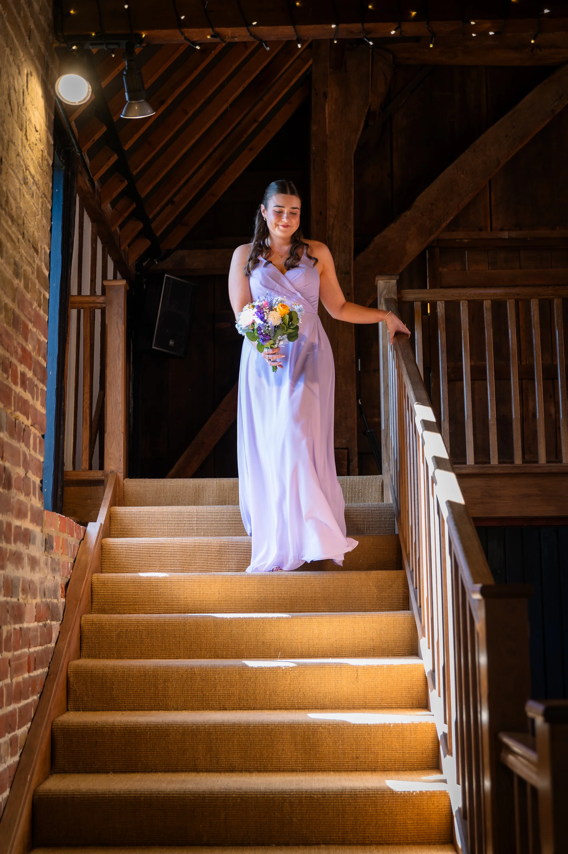 A bridesmaid wearing a light purple dress and holding a colourful bouquet gracefully walks down a wooden carpeted staircase inside a rustic timber-framed barn.