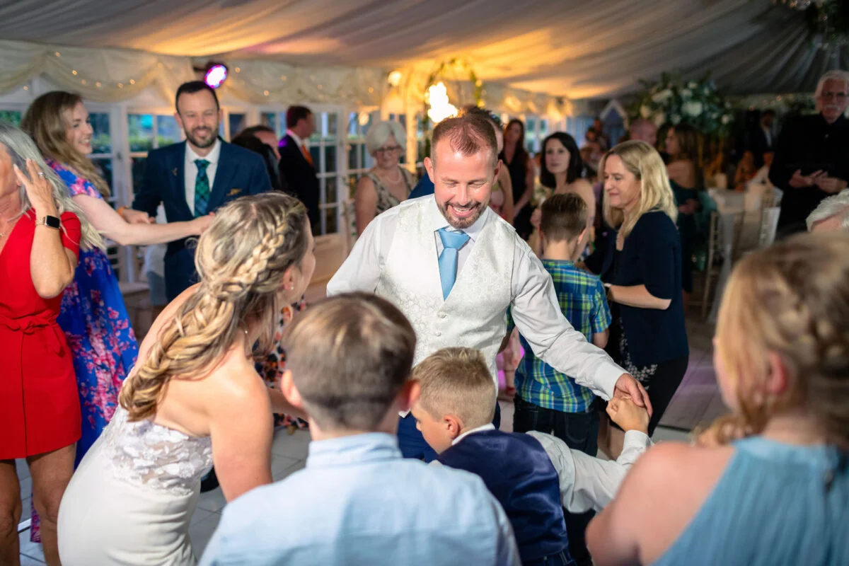 A smiling groom and bride dance joyfully with children and guests on a packed dance floor under a marquee draped with fabric and fairy lights.