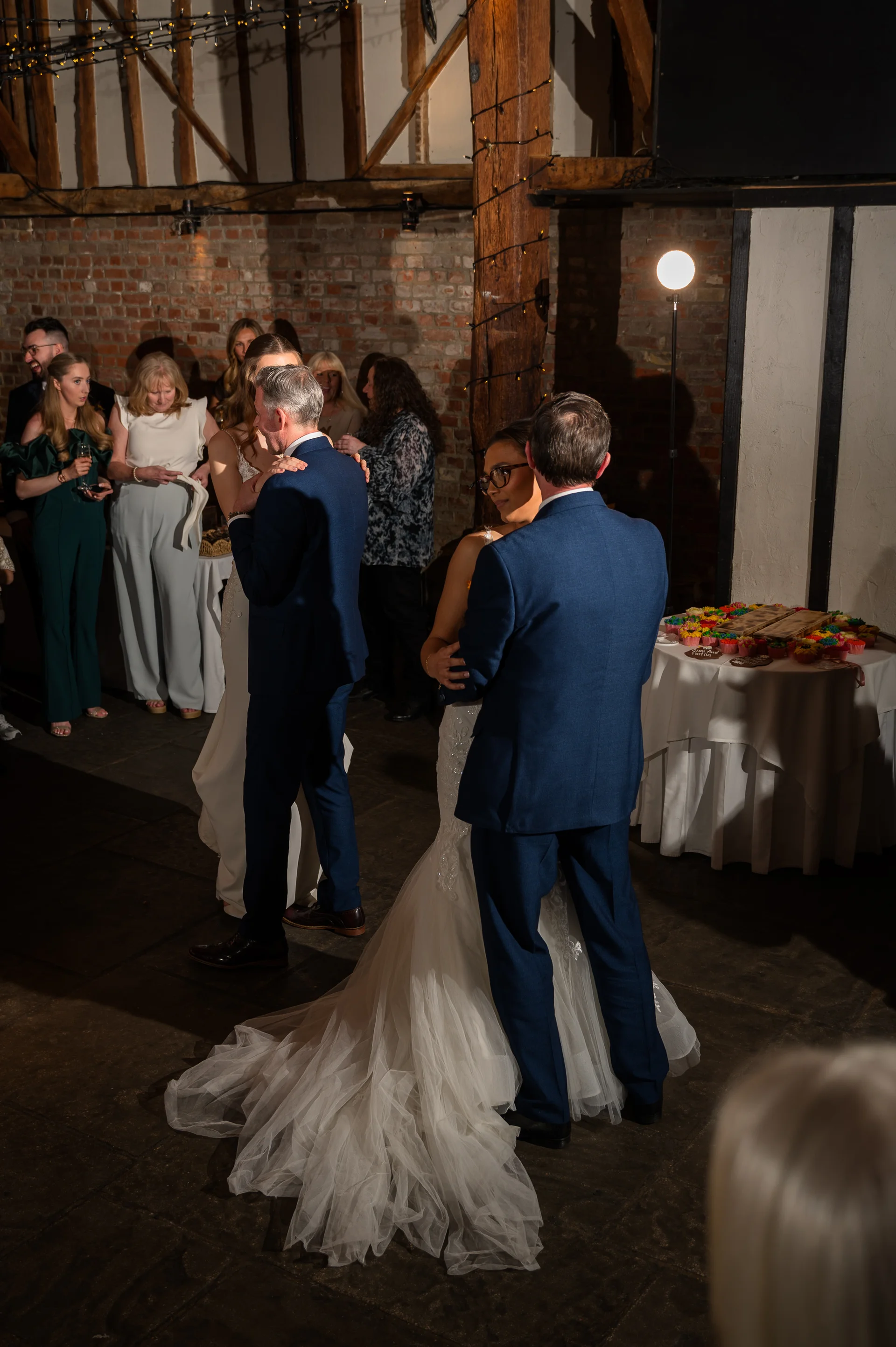 A newlywed couple shares their first dance inside a rustic barn venue with exposed brick walls and warm fairy lighting overhead.