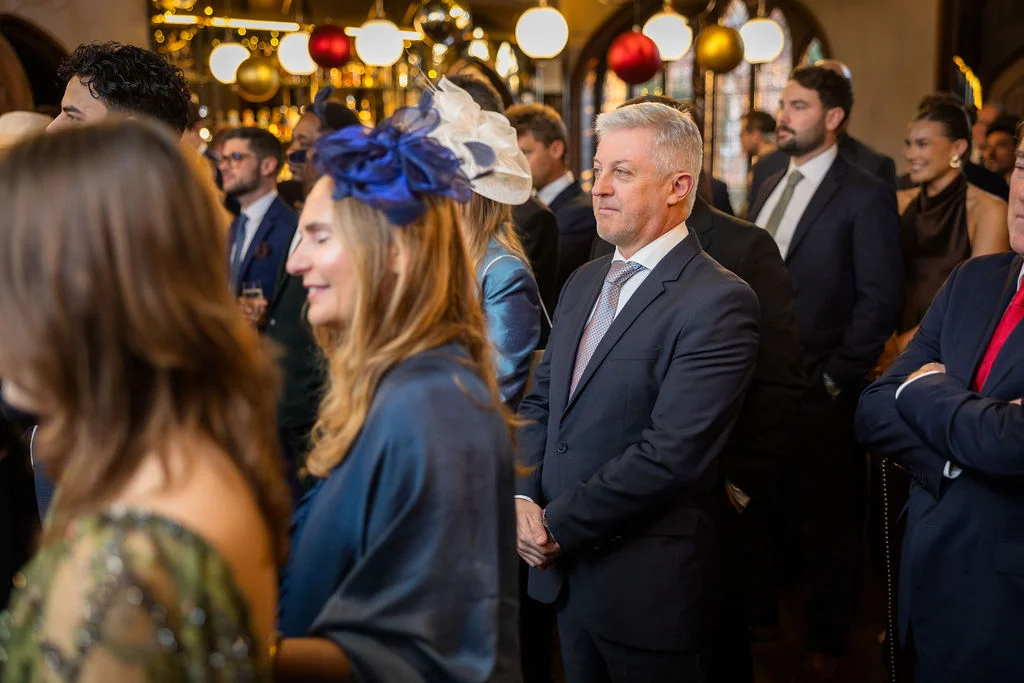 A candid shot of wedding guests gathered indoors, with a mature gentleman in a navy suit watching attentively and a woman in the foreground wearing a blue fascinator.