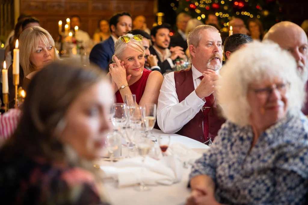 Wedding guests sit around a candlelit table during the wedding breakfast, smiling and listening intently during the speeches.