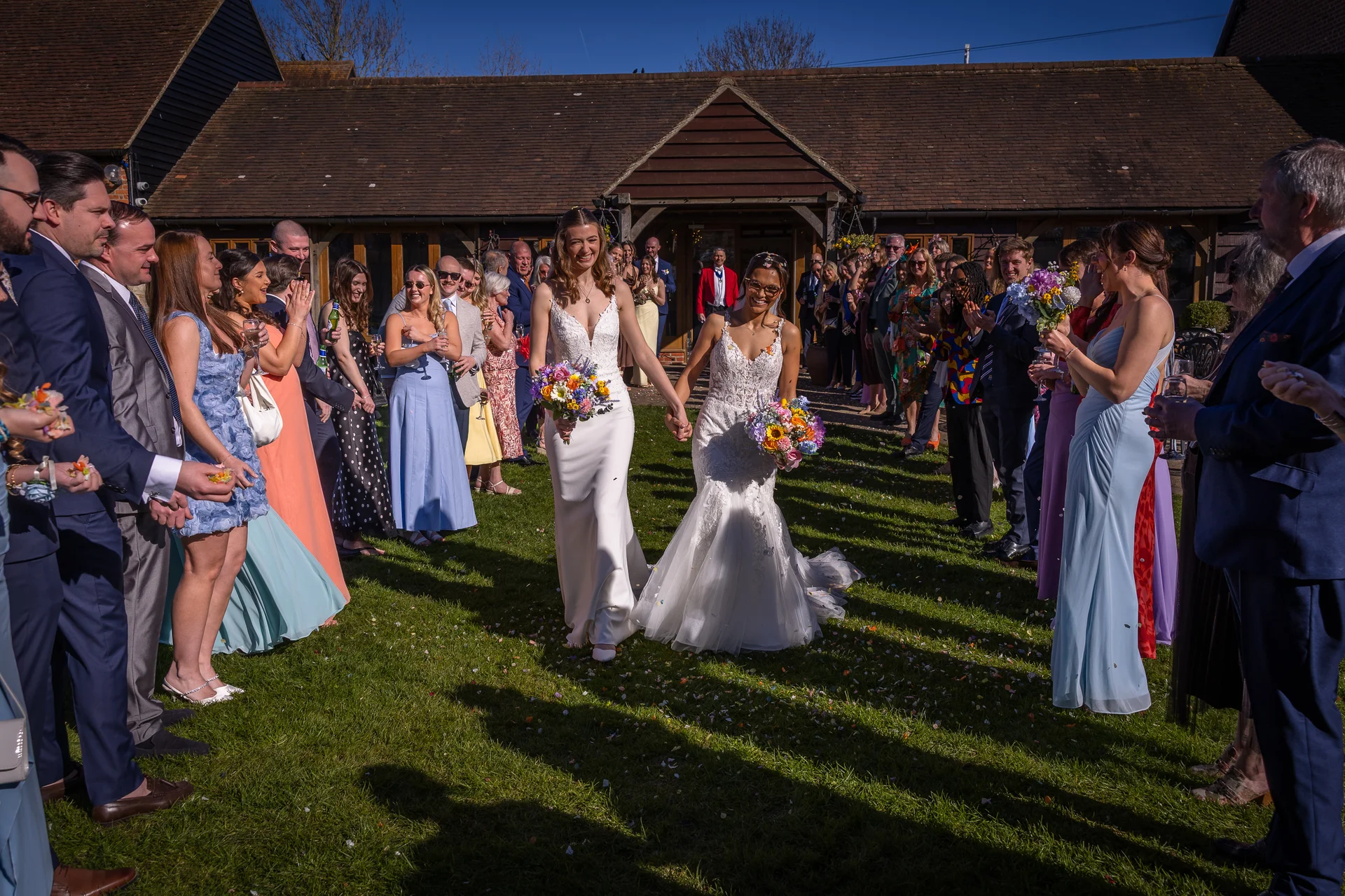 Two smiling brides walk hand in hand down a grassy aisle outside a rustic barn while cheering wedding guests applaud them in the bright sunshine.