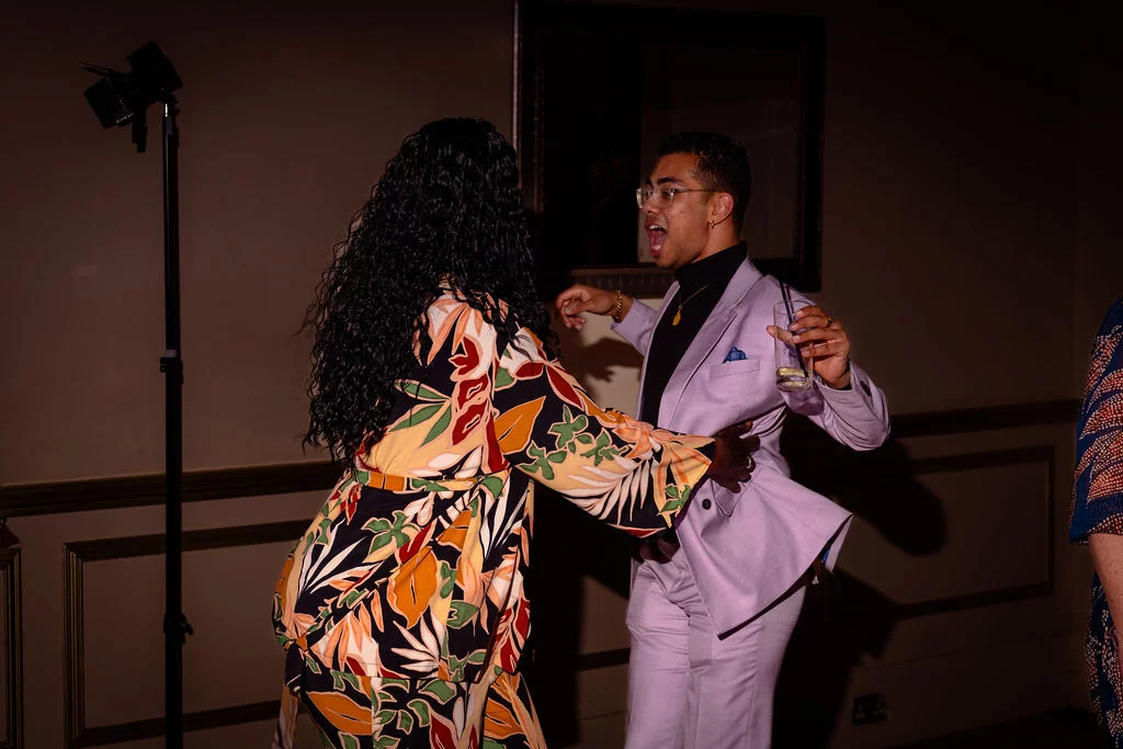 A guest in a vibrant floral outfit dances energetically with a smiling man wearing a light purple suit and turtleneck during an evening reception.