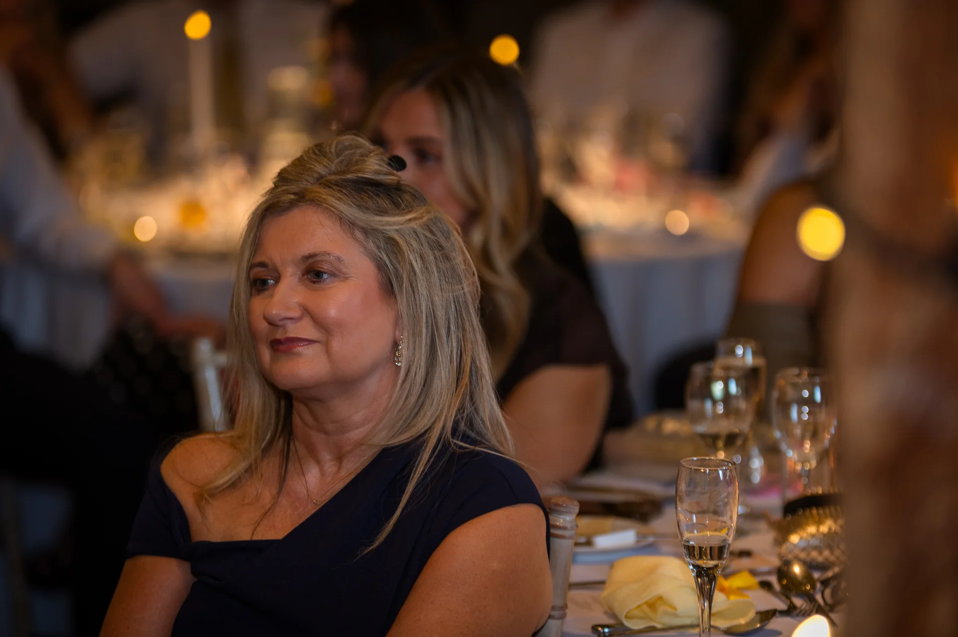 A mature wedding guest smiling during the evening wedding breakfast while seated at a warmly lit reception table.