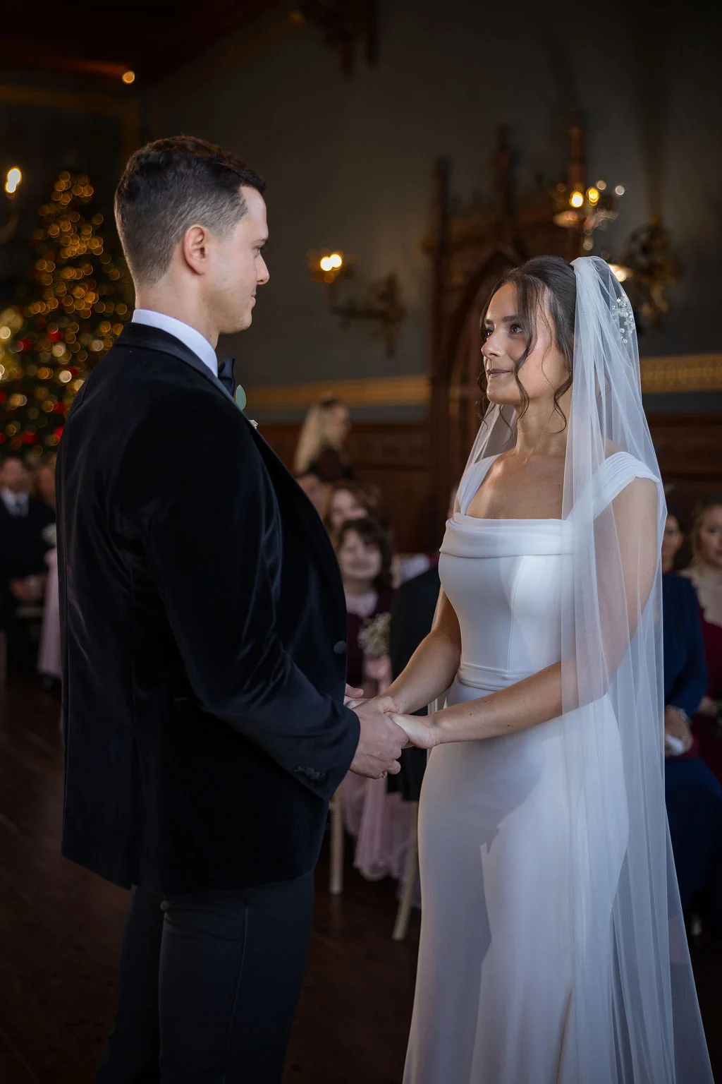 A bride in a white gown and a groom in a black velvet tuxedo hold hands and look at each other during a winter wedding ceremony.