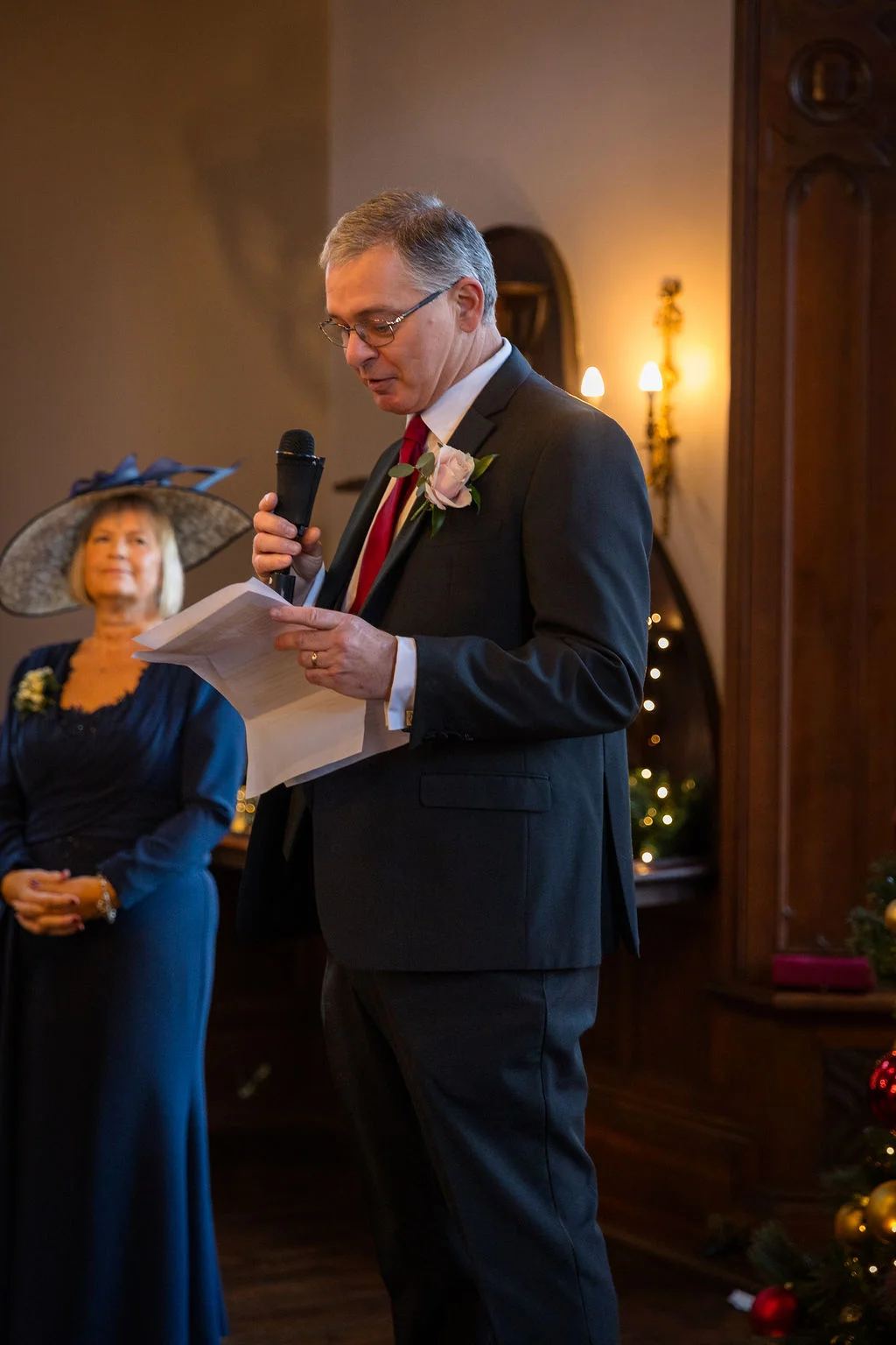 A father of the bride delivers a speech holding notes and a microphone, while the mother of the bride looks on attentively in a dark blue dress and fascinator.