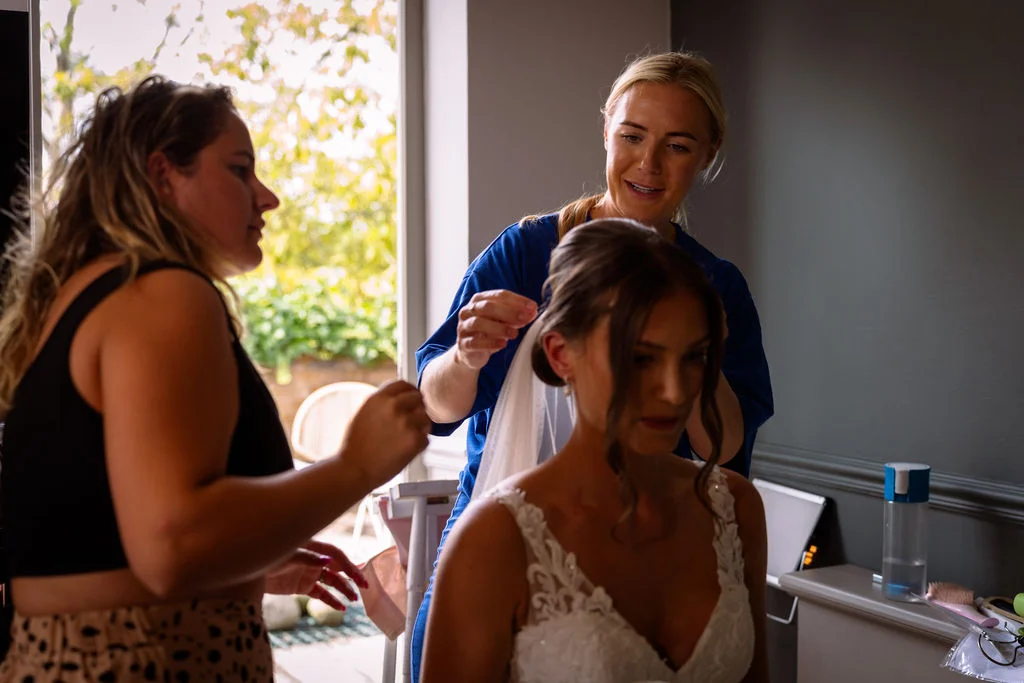 A smiling bride gets her hair styled and veil adjusted by a member of the bridal party in a light-filled room.