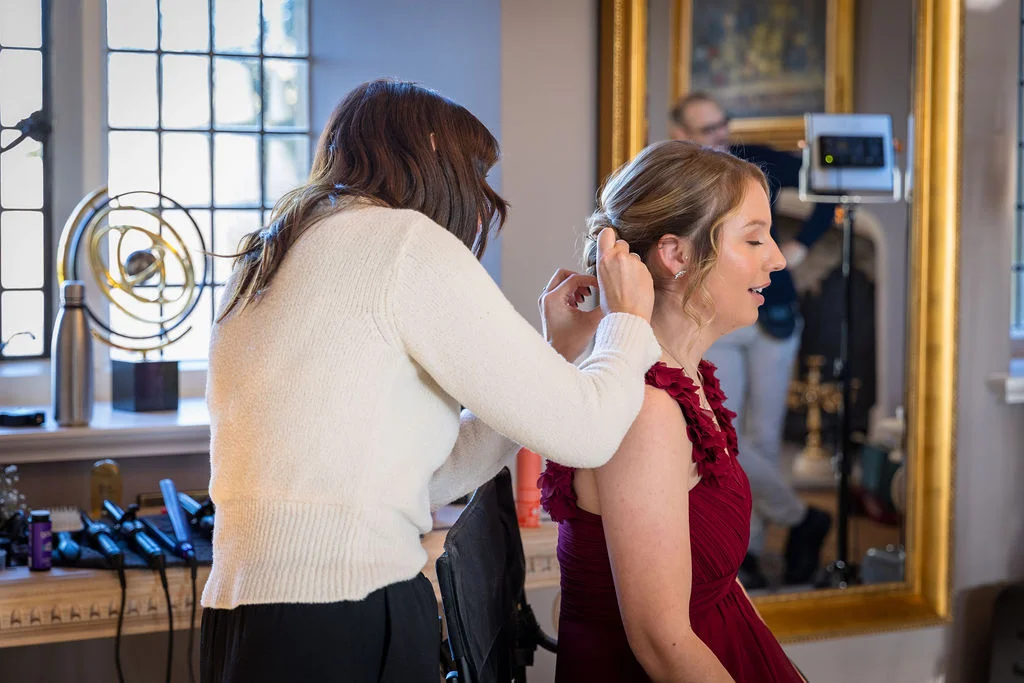 A stylist carefully works on the hair of a bridesmaid wearing a red dress in front of a mirror during morning bridal preparations.
