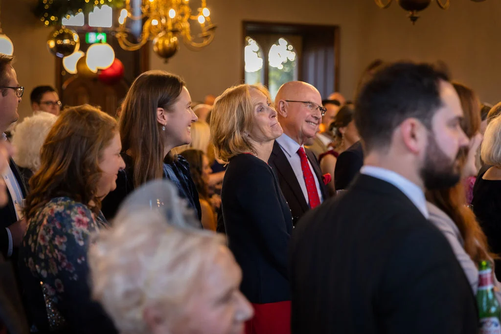 A candid group of wedding guests smiling and laughing while watching speeches inside a venue illuminated by warm chandeliers.