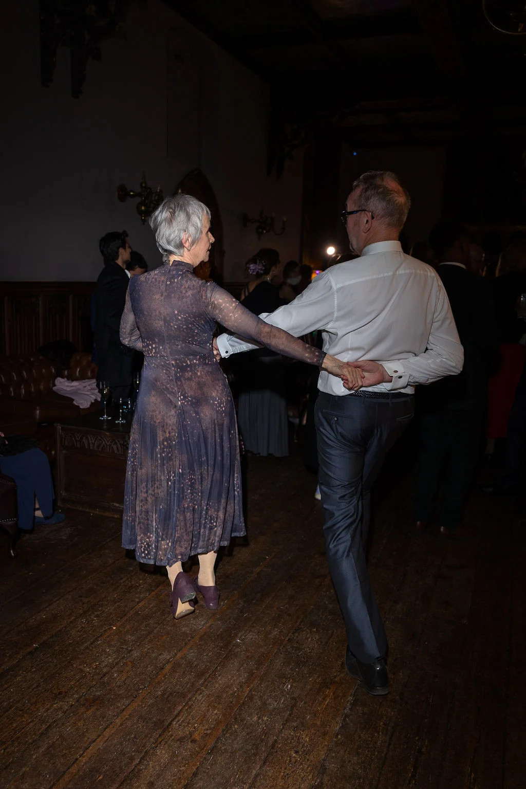An older couple enjoys a dance together on a wooden floor in a dimly lit venue during an evening reception.