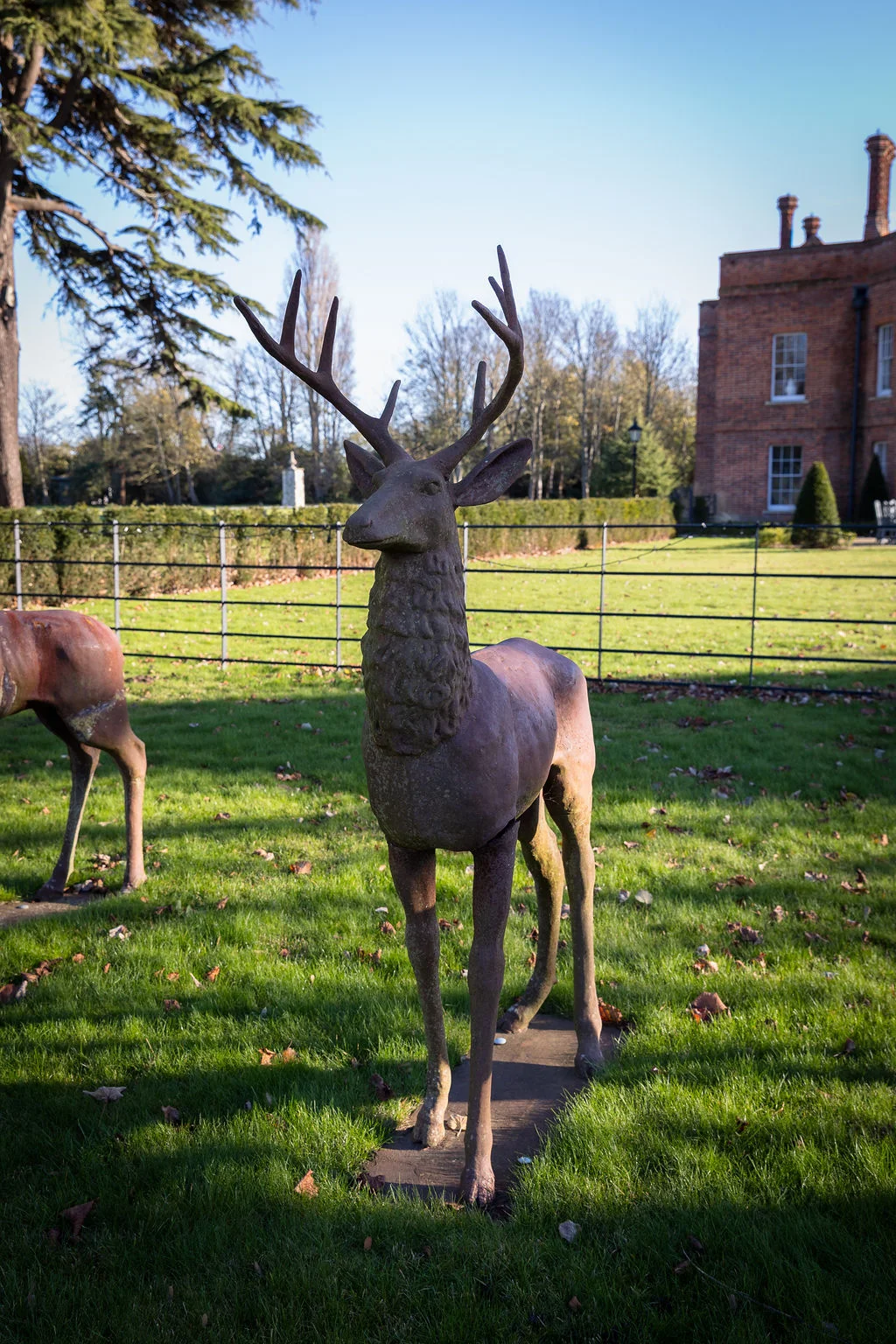 A bronze deer statue stands on a manicured lawn outside a red brick country house on a sunny afternoon.