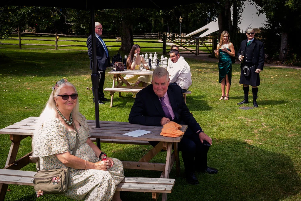 Wedding guests relax on wooden picnic tables in a sunny lawn setting while a man in a traditional kilt and sunglasses stands nearby.