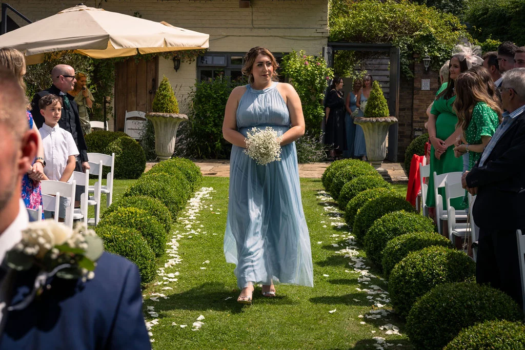 A bridesmaid in a dusty blue chiffon halter-neck dress and holding a baby's breath bouquet walks down an outdoor aisle flanked by topiary and seated guests.