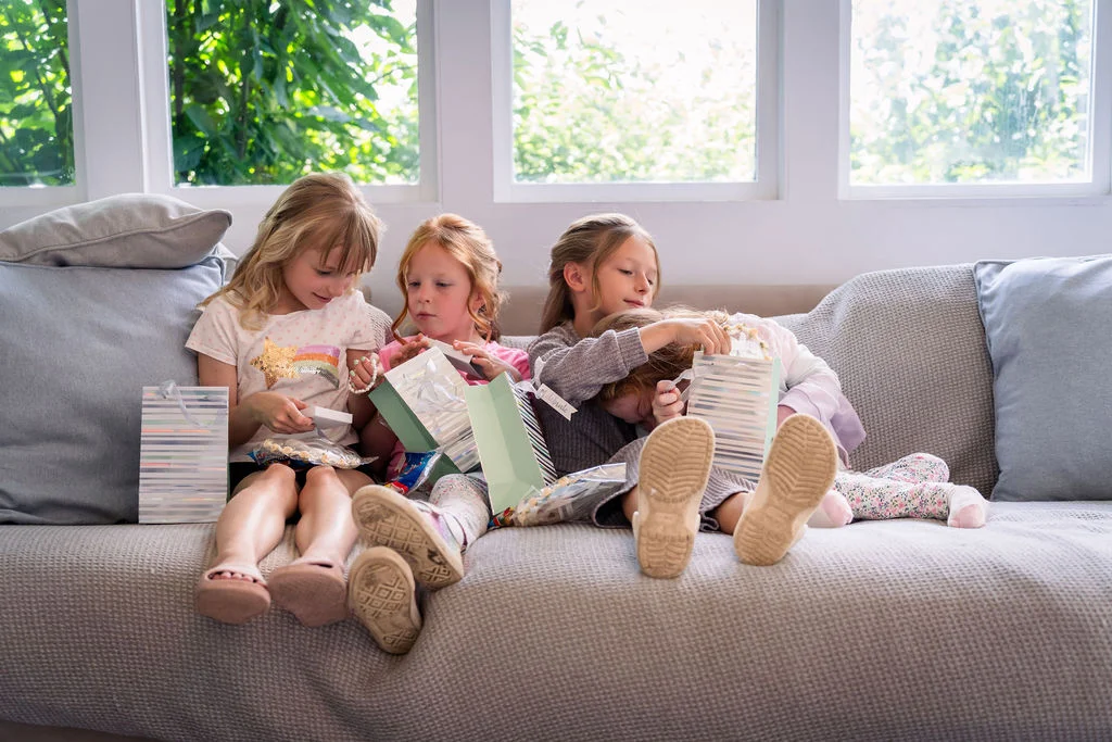 Four young flower girls sit together on a large grey sofa, happily opening gift bags in soft natural light by a window.