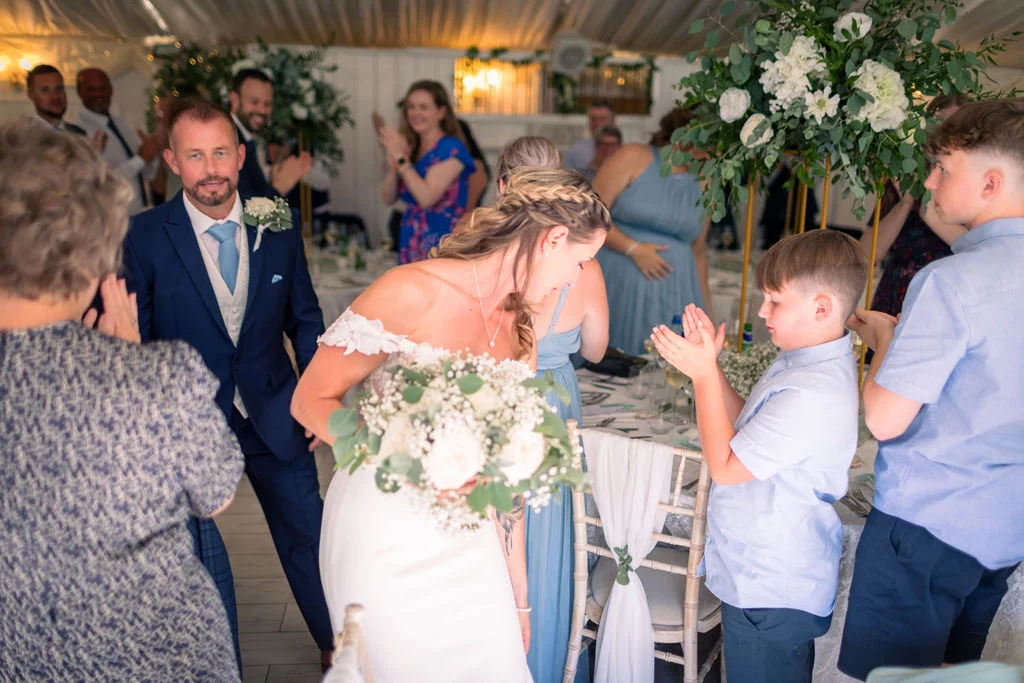 A smiling bride holding a lush white floral bouquet enters the marquee reception while guests applaud enthusiastically.