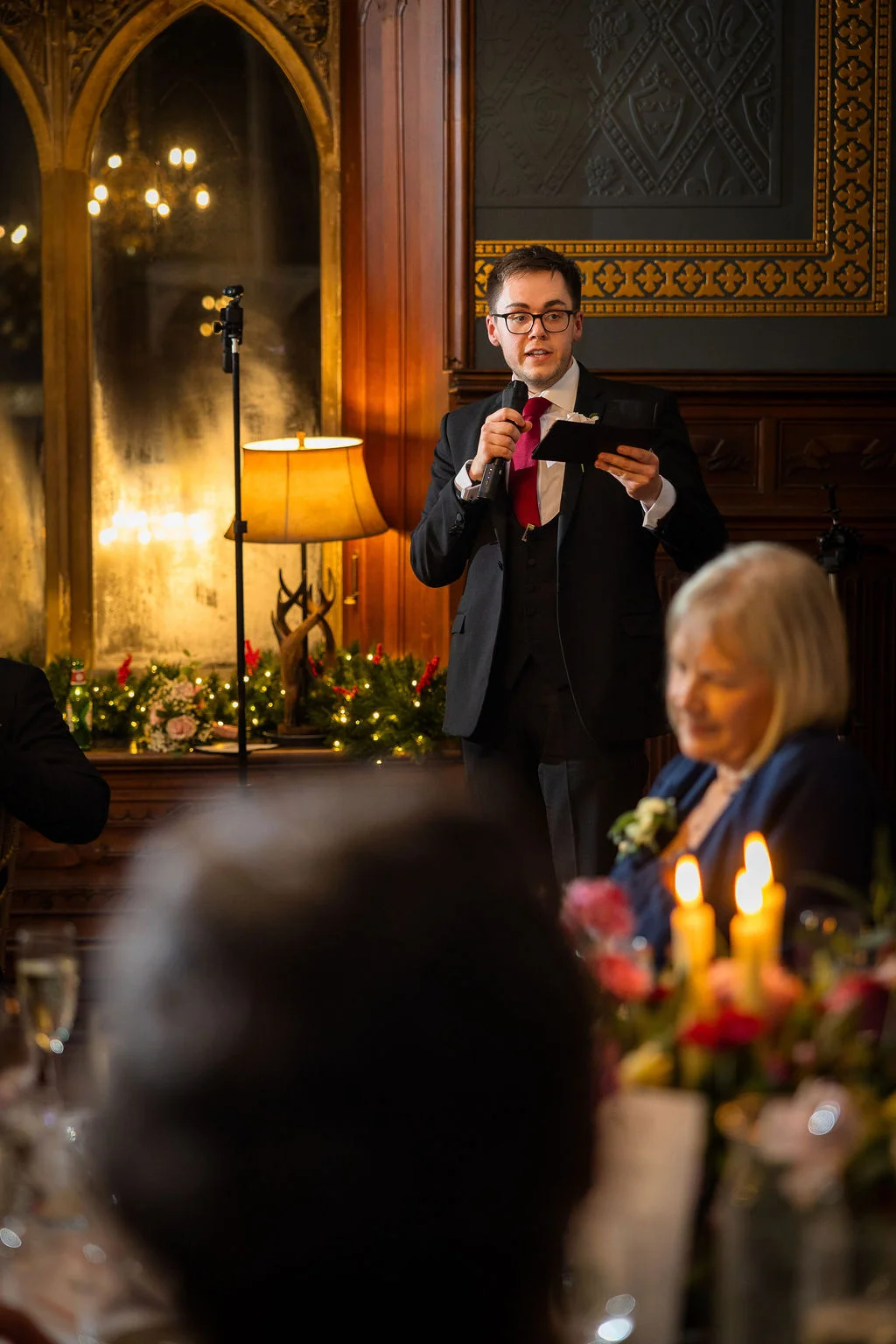 A best man wearing glasses and a three-piece suit delivers a speech during the wedding reception in a wood-panelled room illuminated by candlelight.