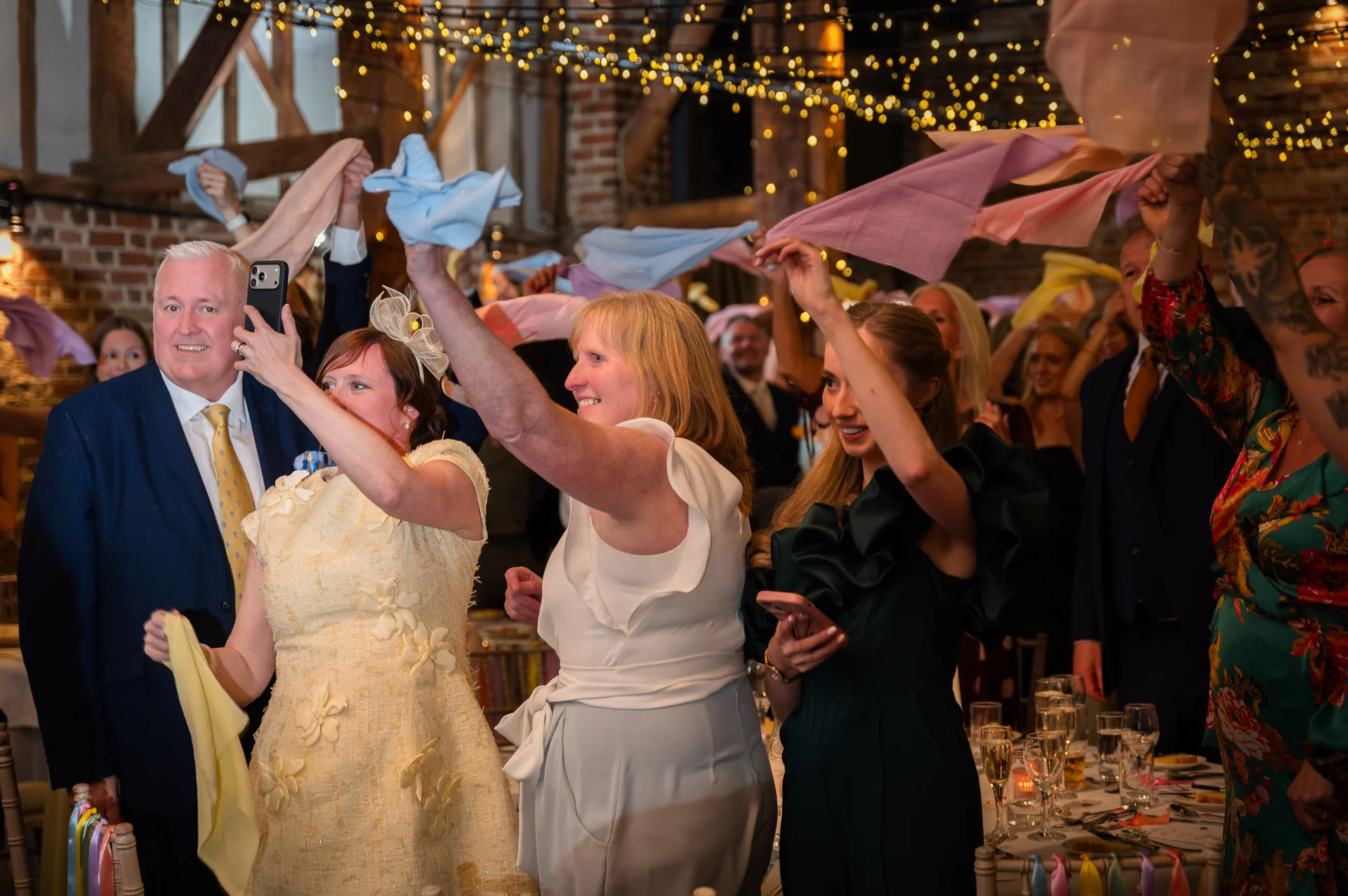 Wedding guests enthusiastically wave napkins in the air during the reception speeches inside a rustic barn illuminated by fairy lights.