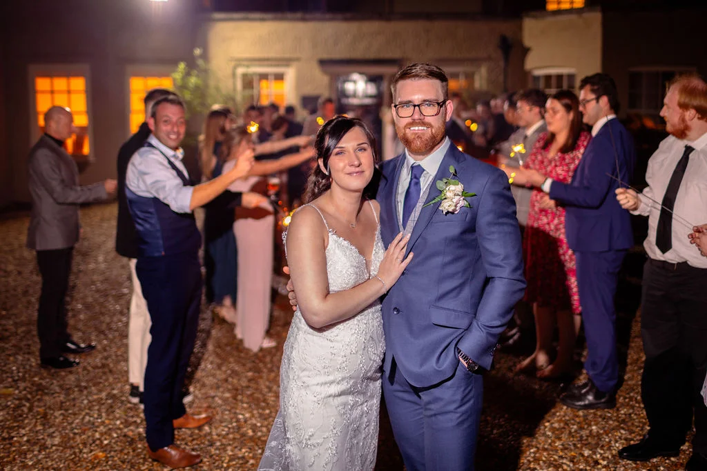 Newlyweds stand smiling in a gravel courtyard at night, framed by wedding guests holding lit sparklers in the background.